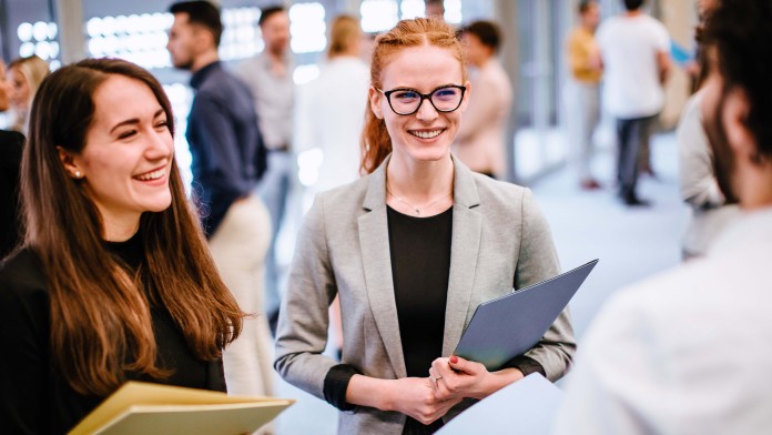 Zwei junge Frauen mit Mappen in der Hand unterhalten sich auf einer Messe mit einem Mann