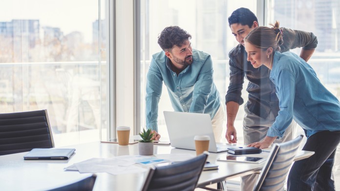 Three young people stand at a conference table and look at a screen