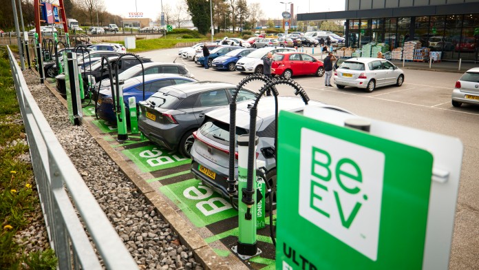 E-Ladestation vollbesetzt mit Autos auf einem Supermarktparkplatz. Im Vordergrund Logo Be.EV