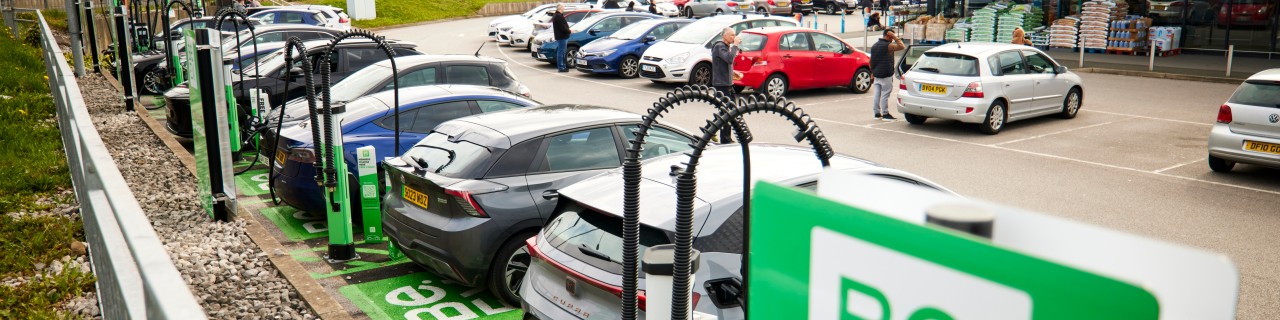 E-Ladestation vollbesetzt mit Autos auf einem Supermarktparkplatz. Im Vordergrund Logo Be.EV