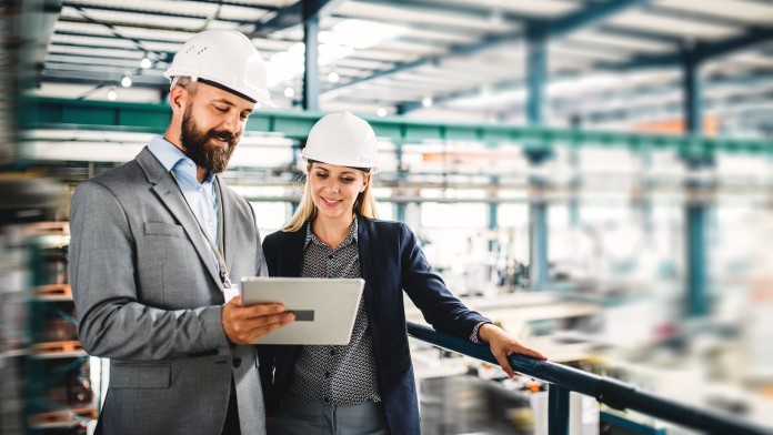 Zwei Menschen in Bürokleidung und mit Helm schauen in ener Werkshalle auf ein Tablet